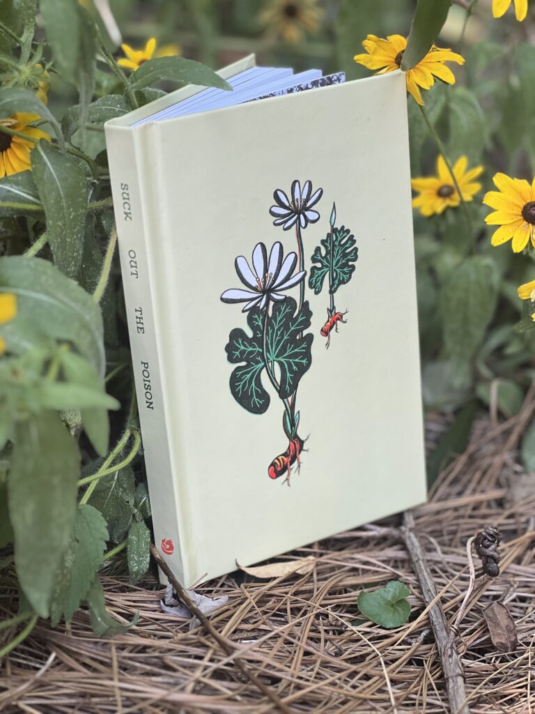 A cream-colored hardcover journal with a bloodroot illustration on the cover and "suck out the poison" on the spine sits in a patch of brown-eyed susans